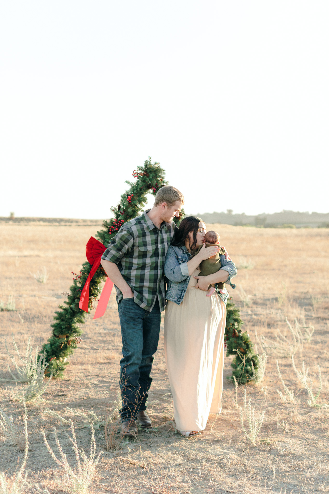 family photos in an open filed with a Christmas garland arch. Mother kisses the baby in her arms while the father watches 